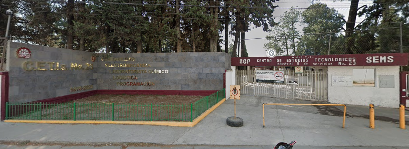 Fotografía en la que se muestra la entrada del plantel conformada por un muro de color gris que tiene unas letras doradas con el nombre del plantel y una reja blanca que da acceso a las instalaciones. Frente al muro hay una jardinera.